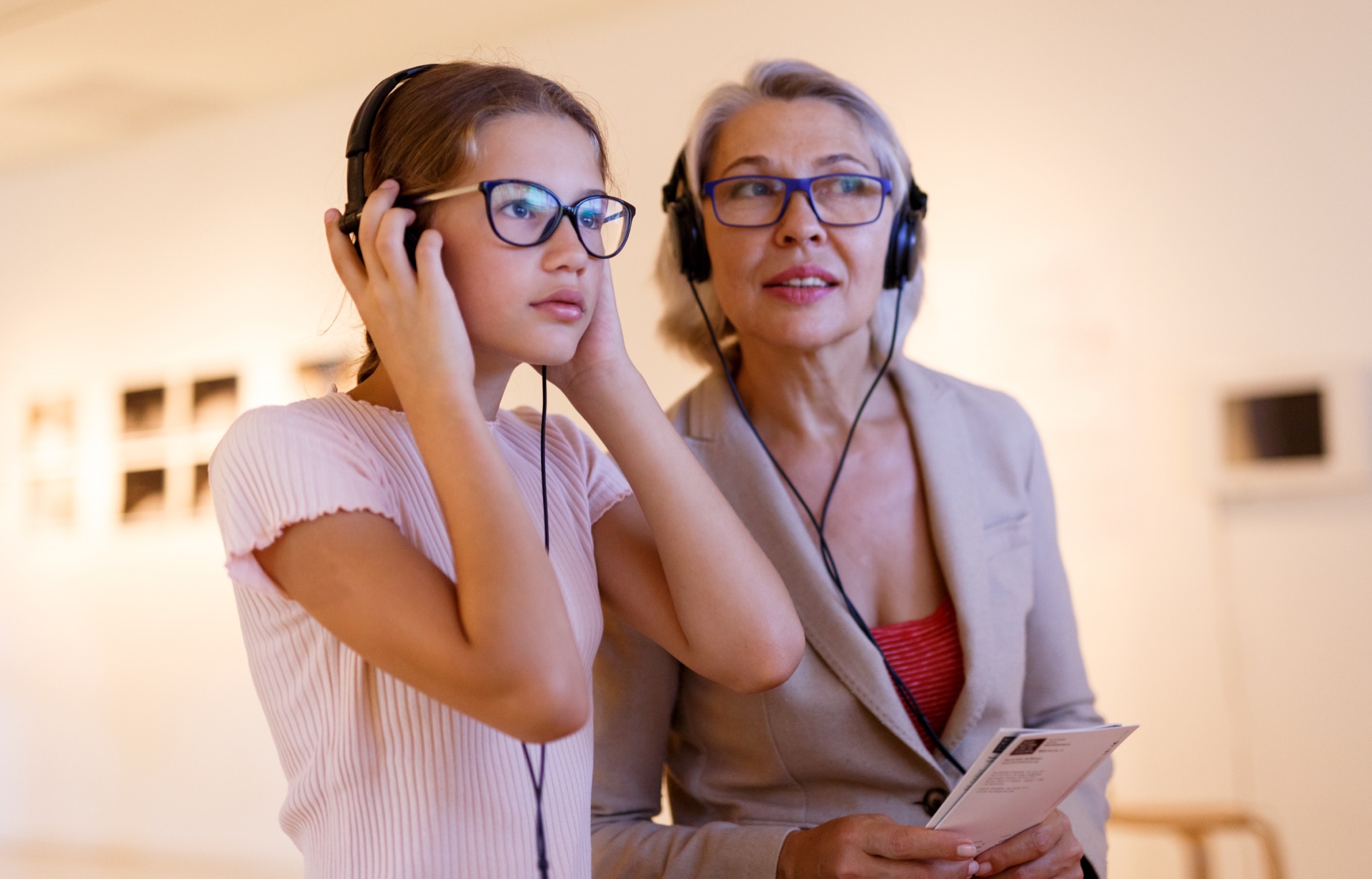 A girl and her grandmother in a gallery, listening to an audio guide on headphones, one of them holding a printed gallery guide.