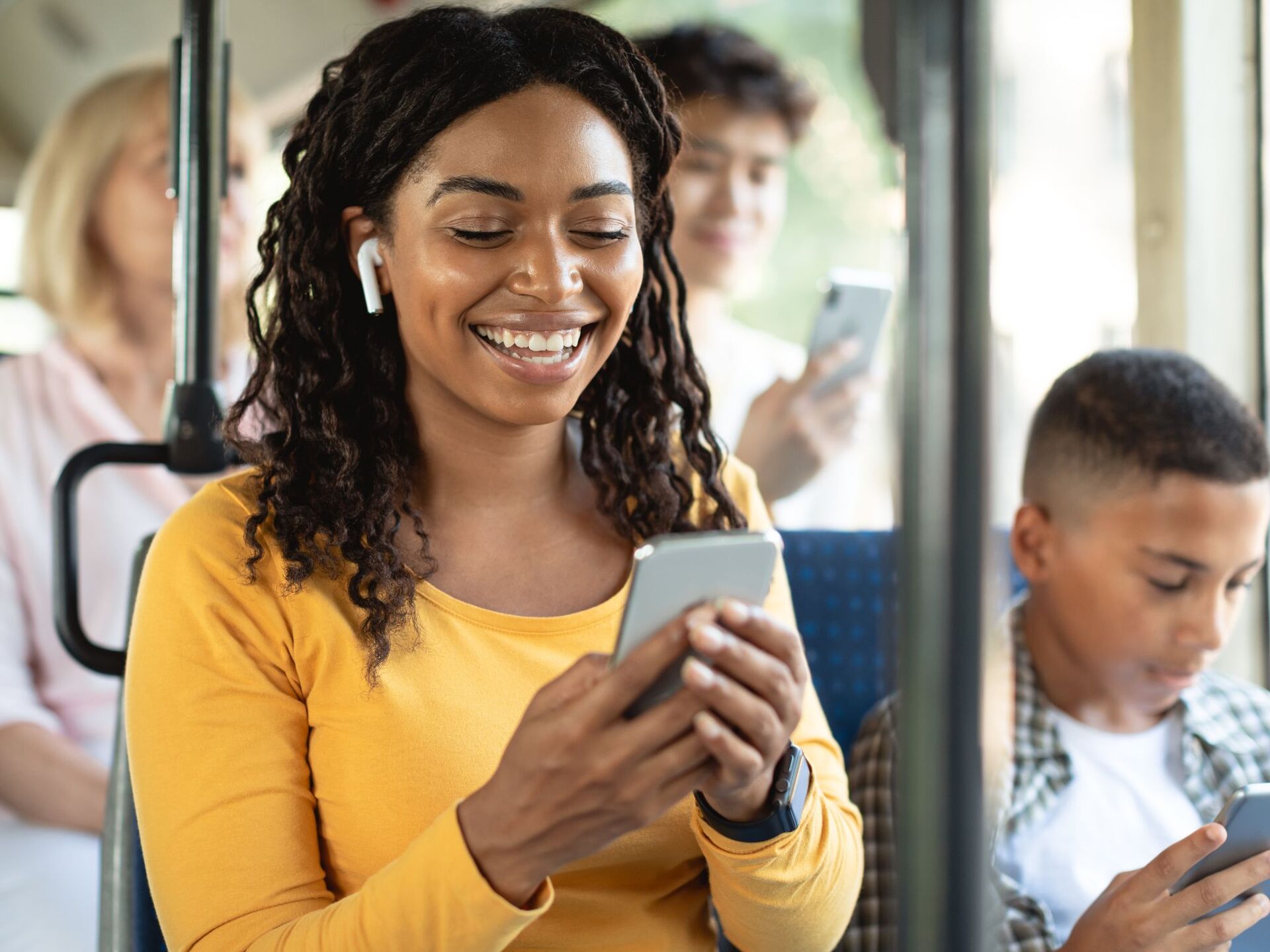 A smiling passenger on a bus, wearing earbuds and looking at her phone, with other passengers on their phones around her.