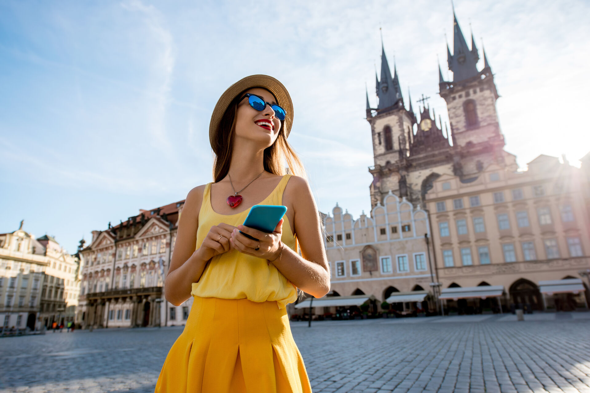 A visitor in a sun hat, holding her phone, in a historic old-town square.